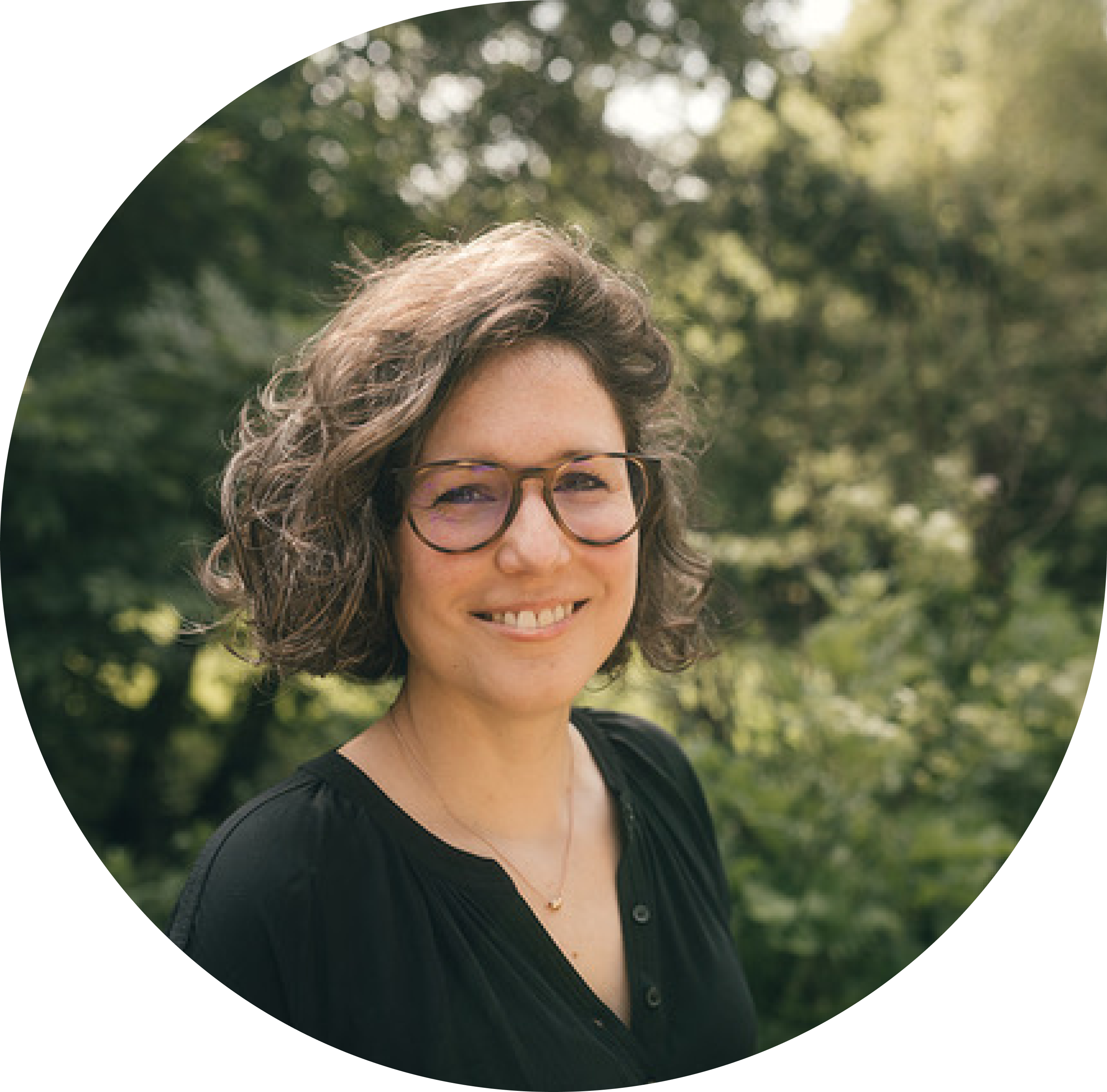 A smiling European woman with glasses, stands in a sunny park. She has a warm, approachable demeanor, with wavy hair and a professional yet casual outfit