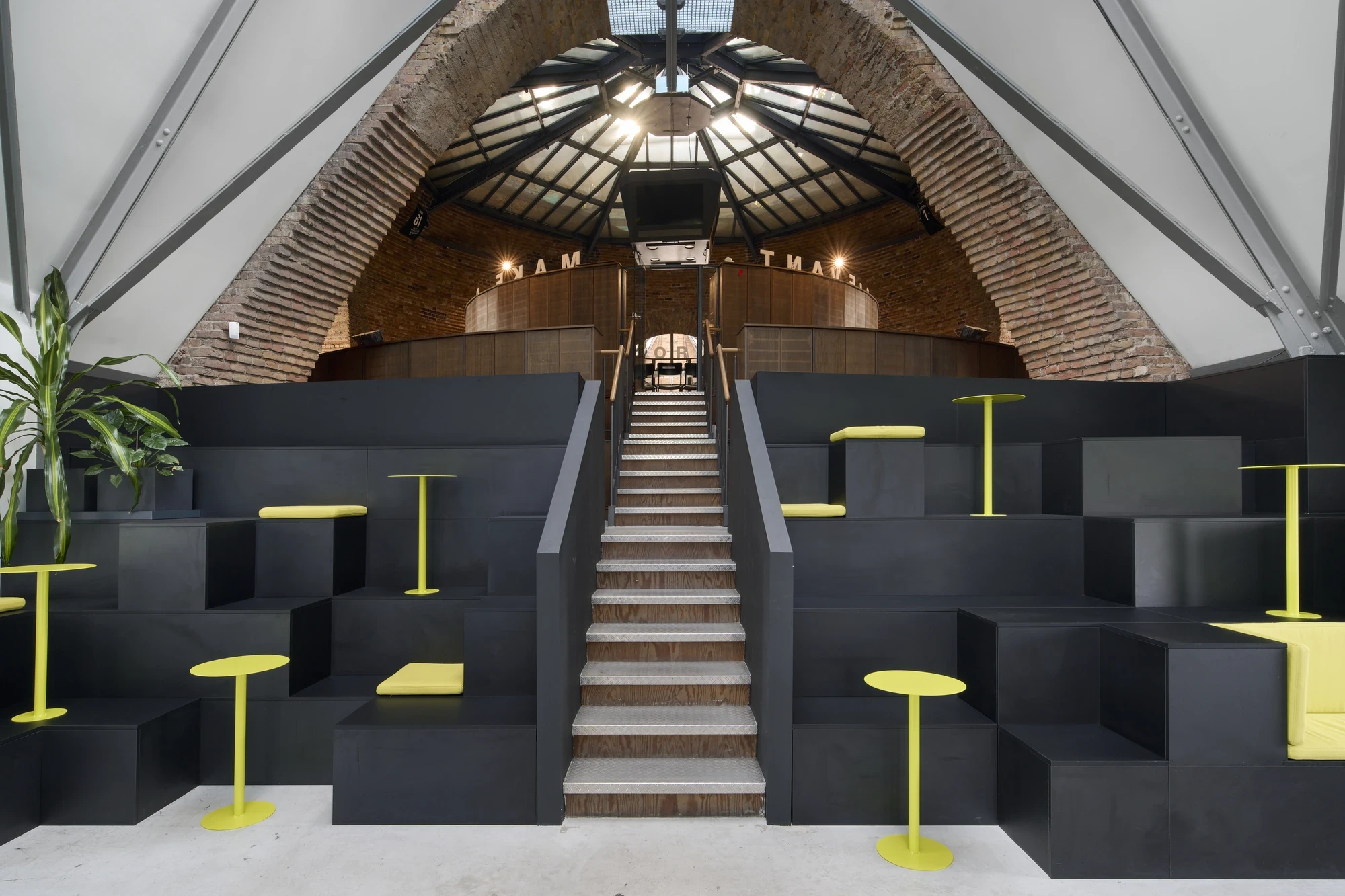Event space inside an arched brick building with central white staircase, black stepped seating platforms and yellow high tables and stools.