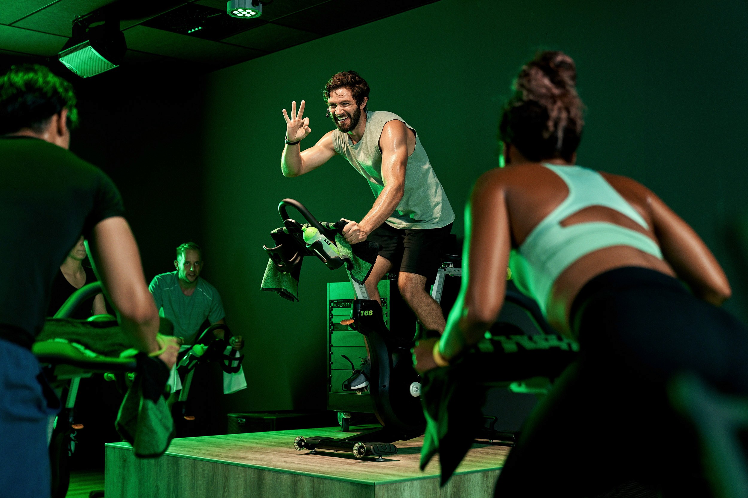 Ein lächelnder Fitnesstrainer leitet eine energiegeladene Spinning-Klasse auf einem Indoor-Bike. Er hebt eine Hand zum Gruß, während zwei Teilnehmer ihm aufmerksam folgen. Die Szene spielt in einem modernen XTRAFIT Fitnessstudio mit grüner Beleuchtung und einem schlichten Hintergrund.