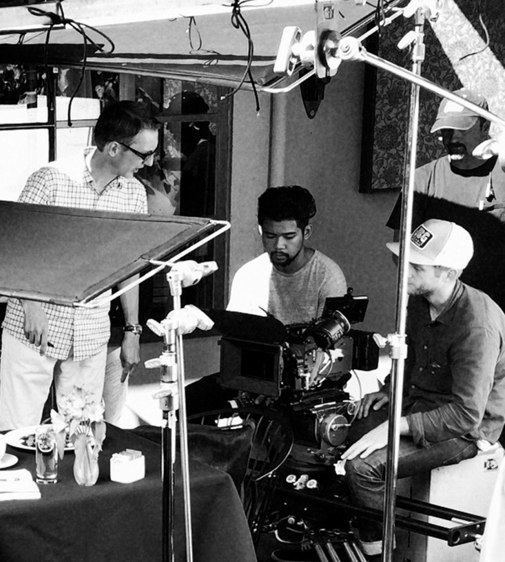 A black-and-white photo captures a live-action production crew on set, gathered around a camera setup at a restaurant table scene. One crew member operates the camera while others review the monitor and discuss the shot. Lighting gear and reflectors frame the foreground, and a table set with flowers and drinks suggests a scripted dining scene in progress. The atmosphere is collaborative and focused.