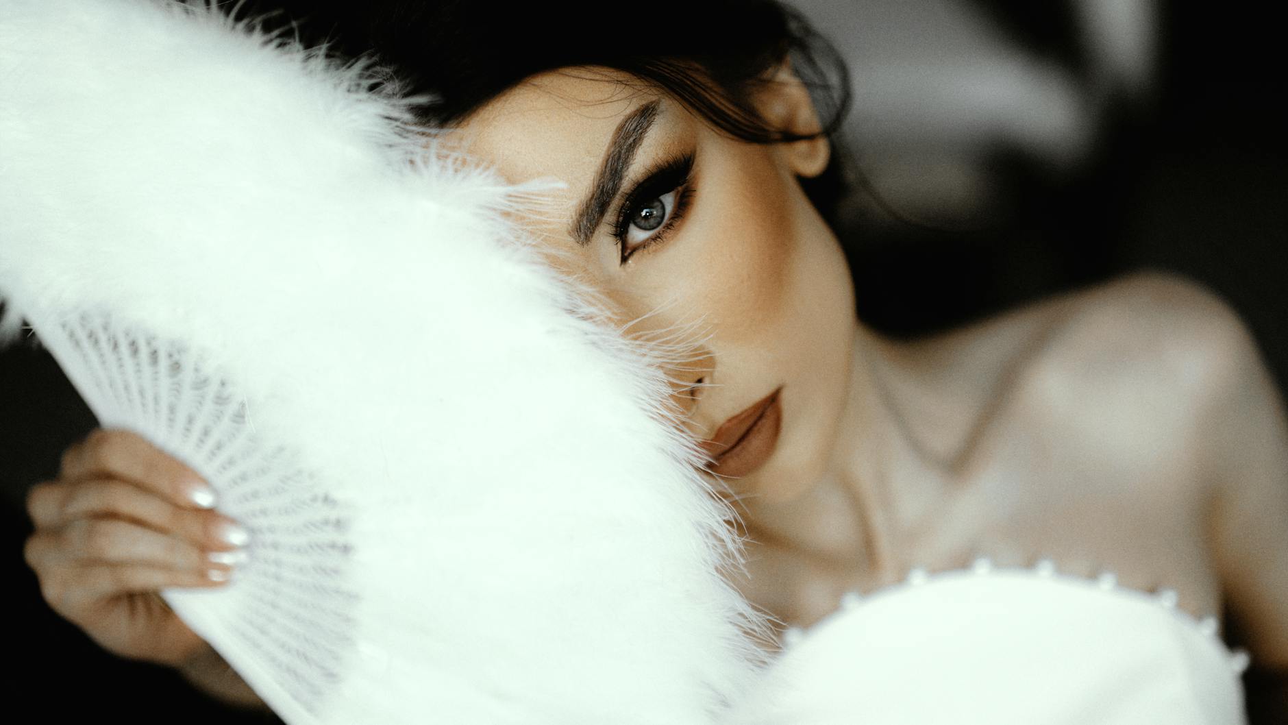 Close-up of a bride's face partially hidden by a feather fan, exuding elegance and mystery.