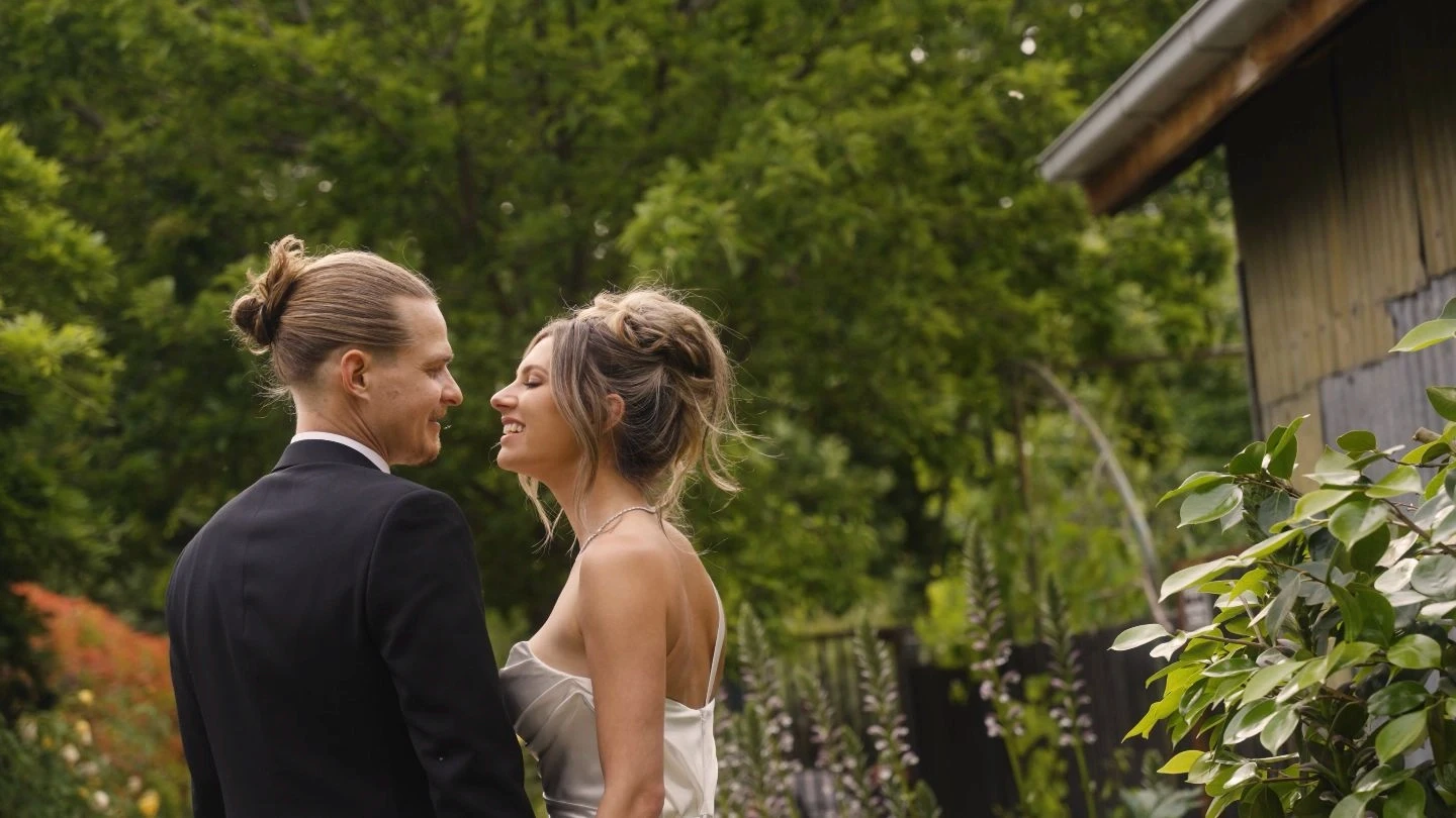 A smiling bride and groom stand closely together outdoors, surrounded by lush green foliage and blooming plants, next to a rustic wooden building, capturing a joyful wedding moment.