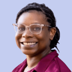 A smiling woman with short dark hair pulled back and clear-framed glasses wearing a burgundy or maroon colored top, photographed against a soft lavender background.