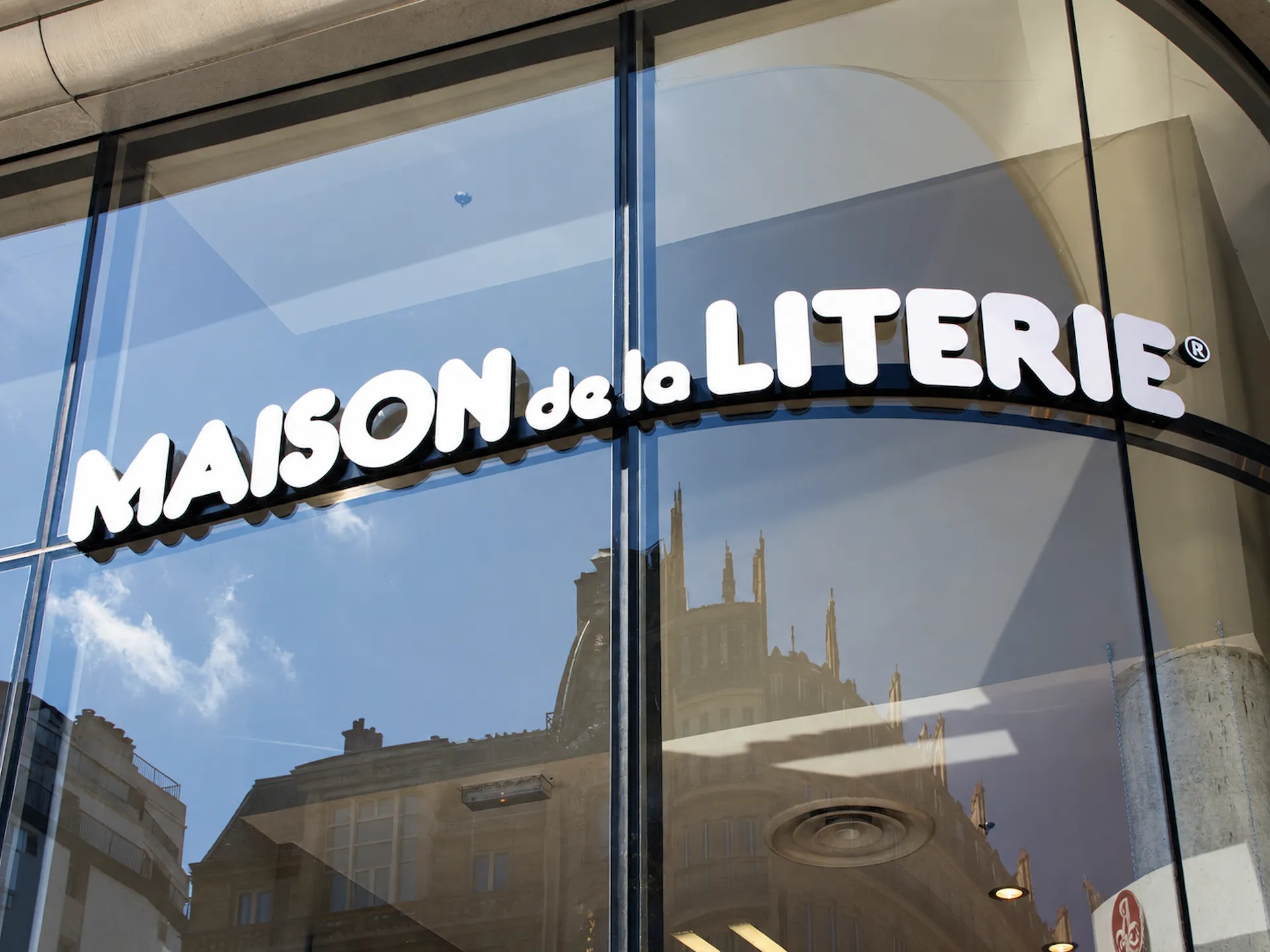 Bílý světelný nápis MAISON de la LITERIE z 3D písmen na zaoblené prosklené fasádě prodejny