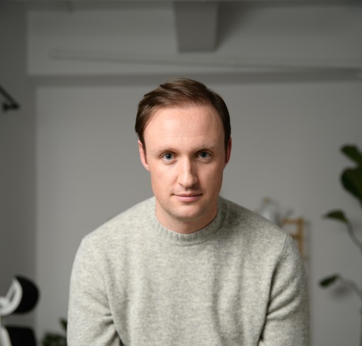 A smiling man with glasses and a dark grey zip-up sweater stands confidently outdoors, embodying PointOne's robust.