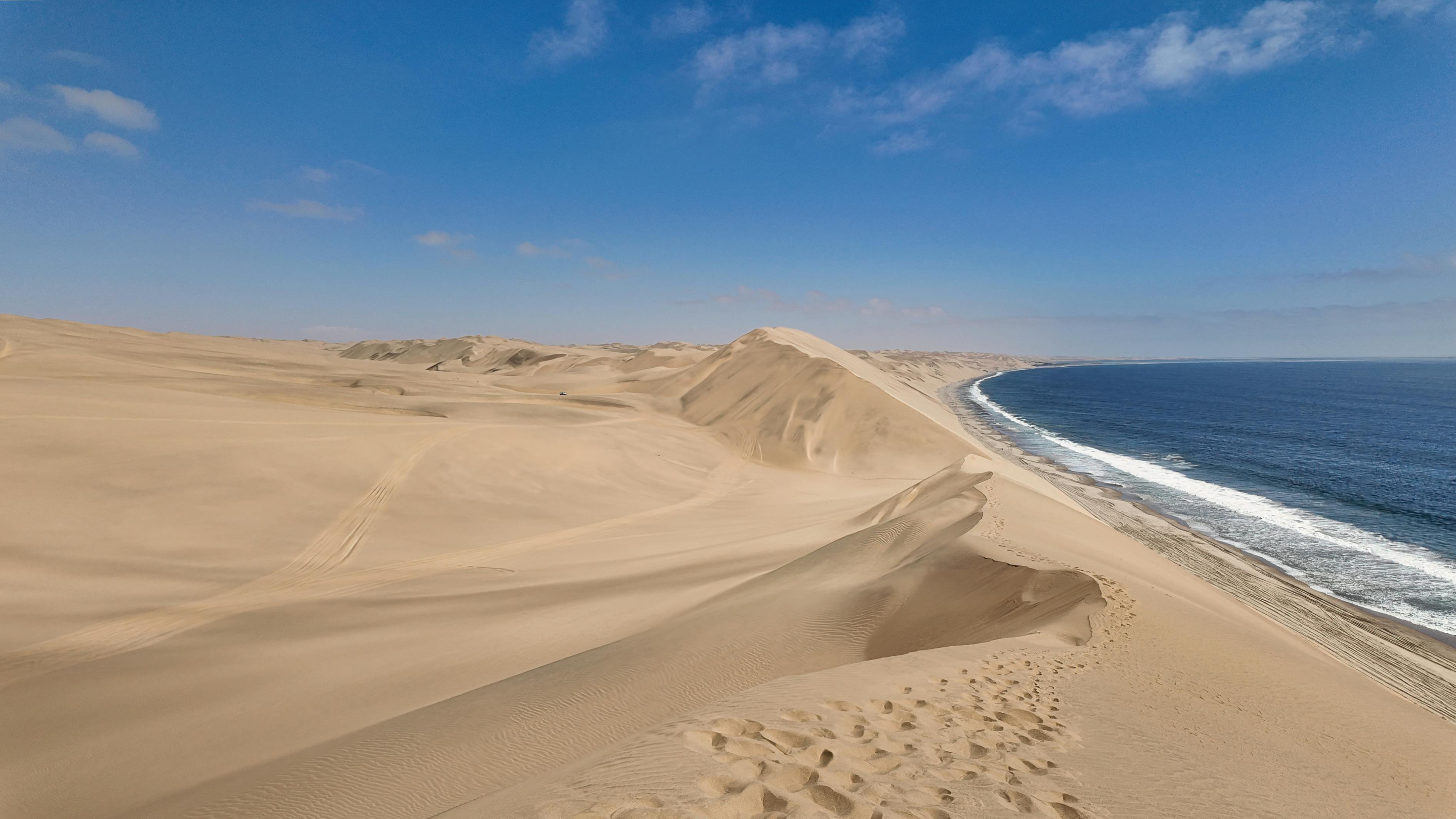 A sandy desert landscape meets the ocean under a clear blue sky, with gentle waves lapping at the shore.