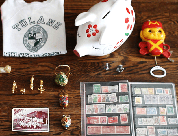 A wooden table displays a collection of items: a white Tulane University sweatshirt, a white piggy bank with red and green floral designs, a yellow and red M&M's character, a small white plastic ring, a green tea kettle, a gold candlestick set, a small gold owl figurine, a small red and white picture frame, a book of postage stamps, and a small white figurine.