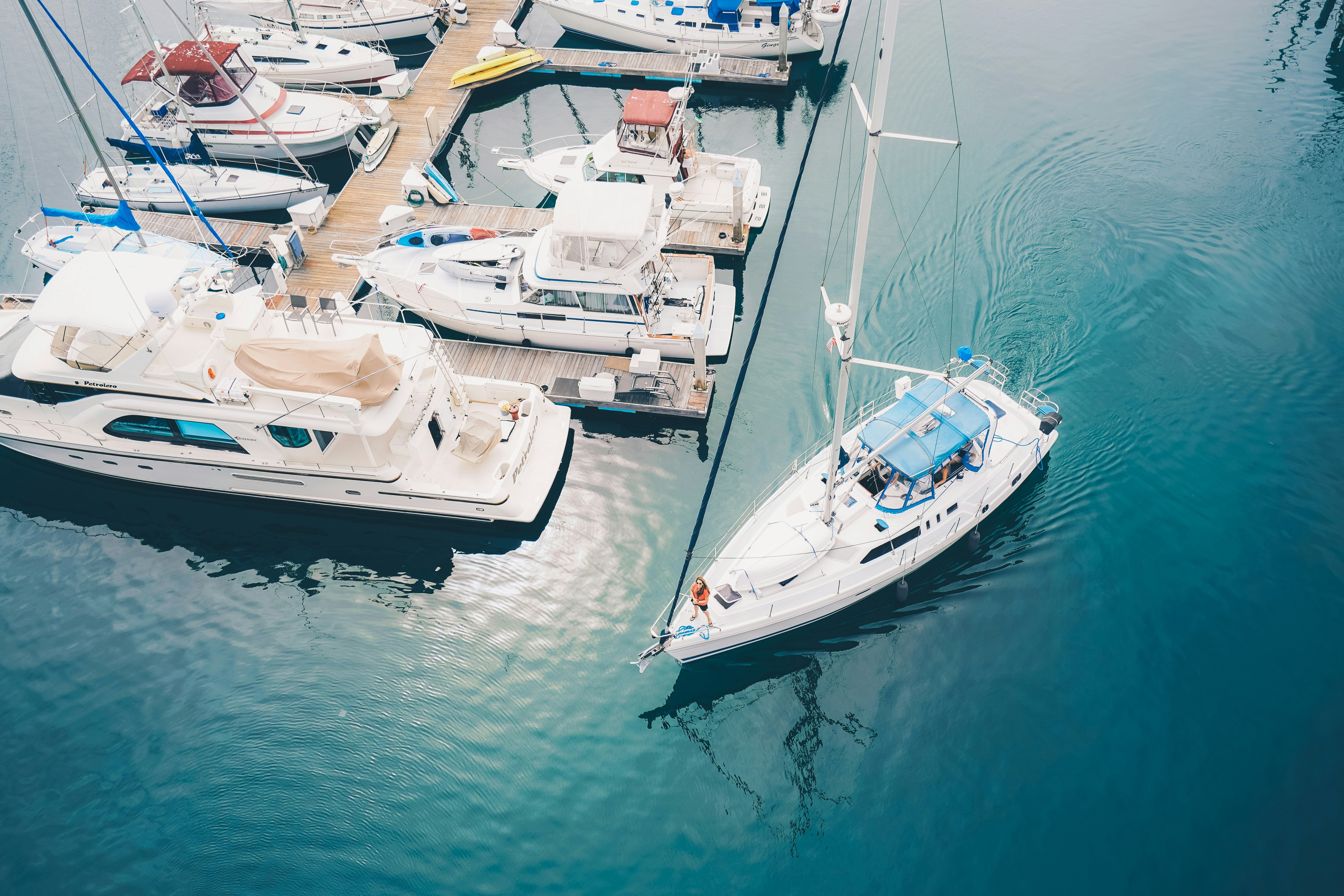 Aerial view of a marina with several yachts and sailboats docked, featuring calm blue waters and one sailboat actively maneuvering towards an open dock space.