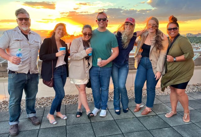 Seven people stand on a rooftop at sunset, smiling with drinks in hand against a cityscape—capturing the vibrant, collaborative environment that defines TBC.