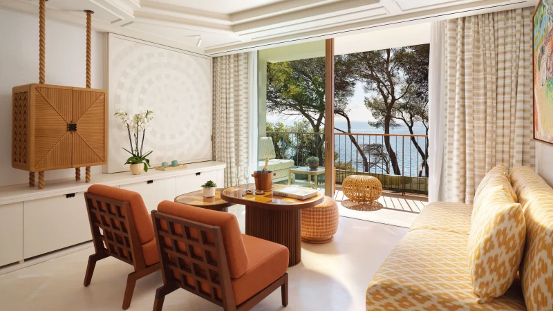 Ein elegantes Wohnzimmer mit zwei orangefarbenen Sesseln, einem Sofa mit gelbem Muster, einem hölzernen Barschrank und einem Balkon mit Blick auf Pinien und das Meer.