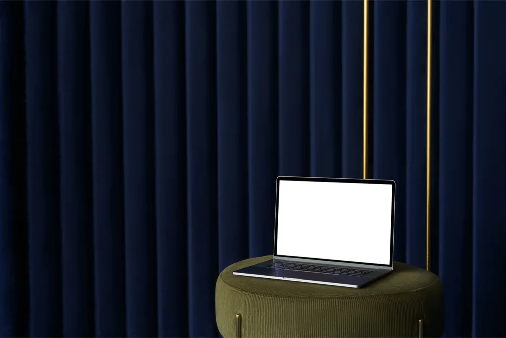 Laptop mockup with a blank screen placed on an olive-green upholstered stool. The background features luxurious deep blue curtains with gold accents, adding a sophisticated touch.