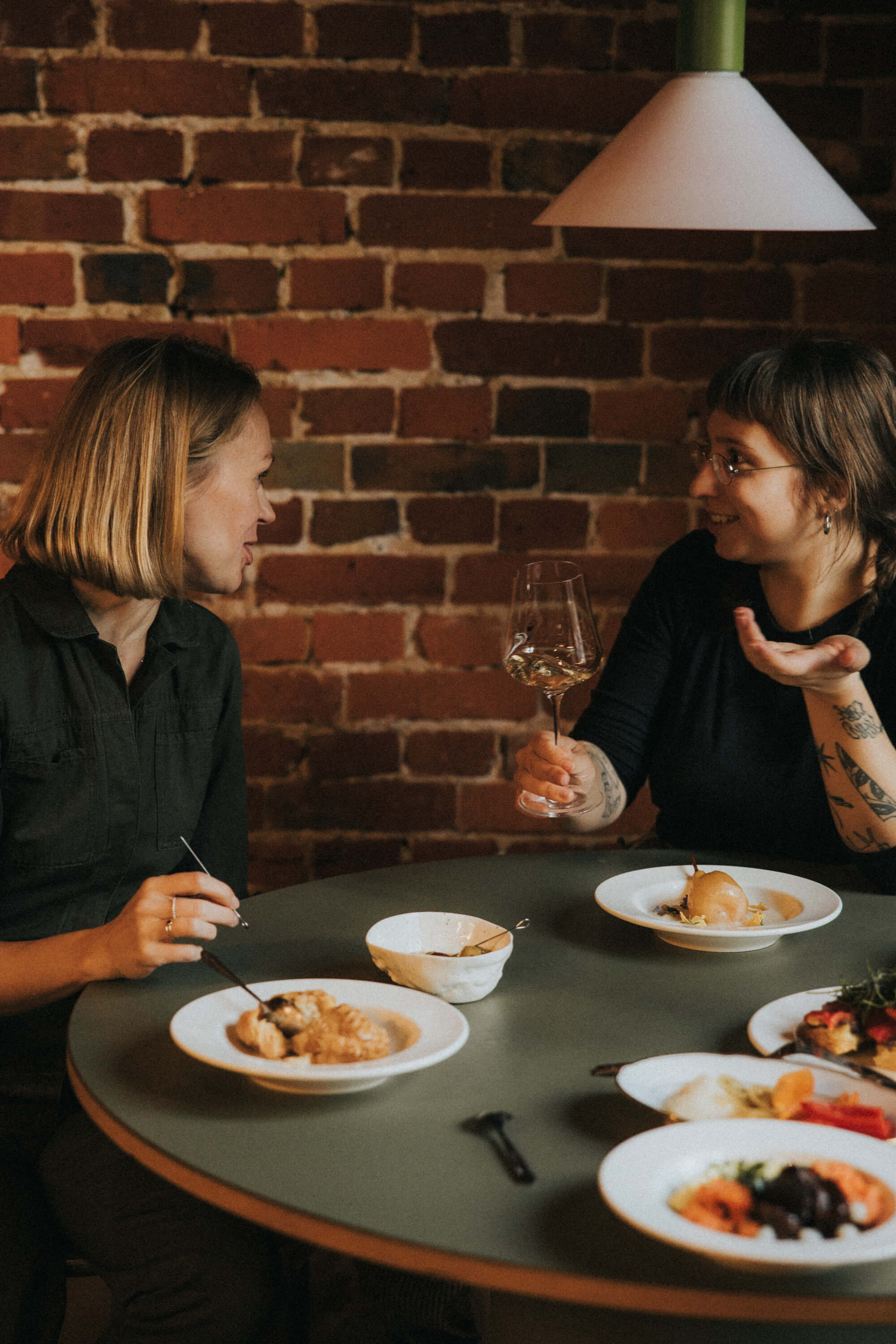 Two people sit at a table conversing, sharing a meal in a cozy setting with brick walls and plates of food.
