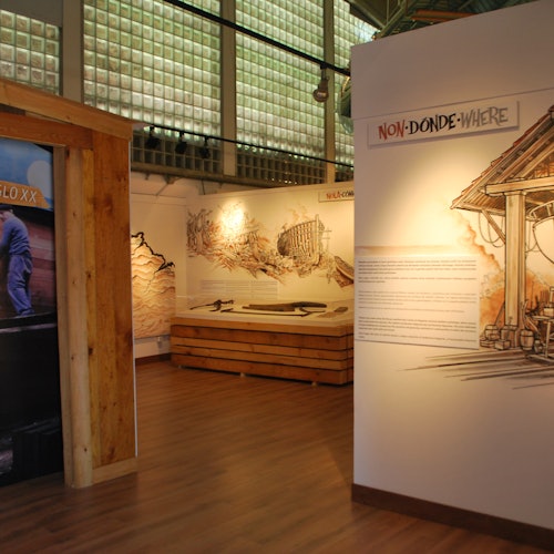 Museum exhibit room featuring illustrations, text panels, and architectural models. Sign reads "NON-DONDE-WHERE" with wooden accents.