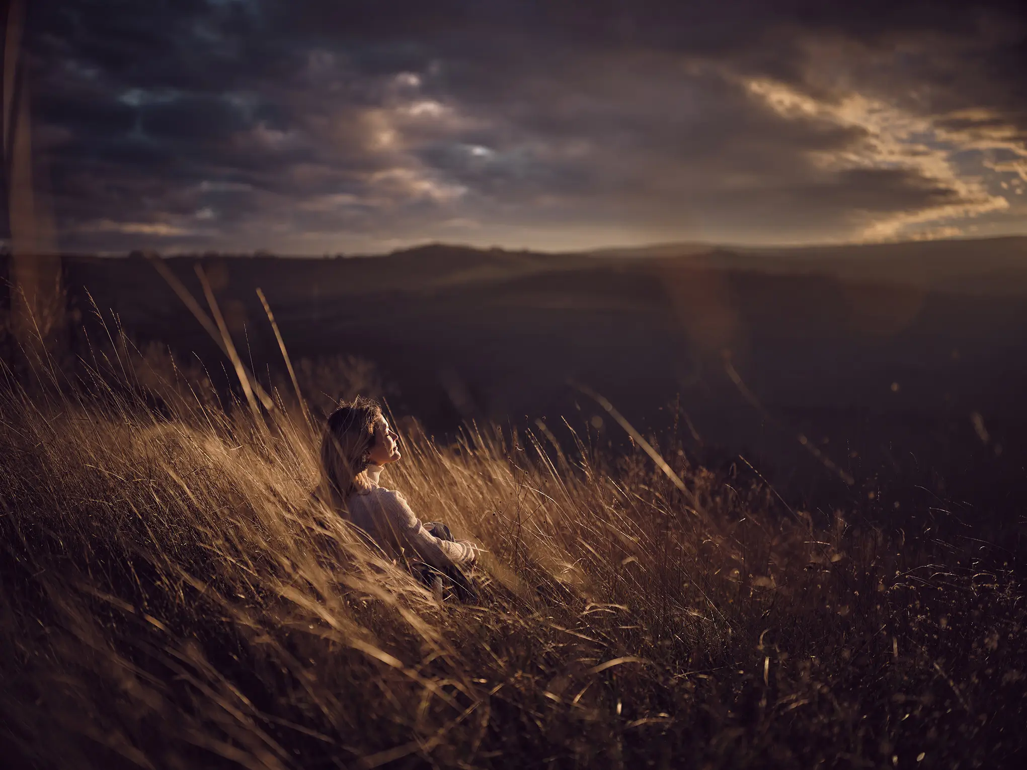 A silhouette stands on a windswept landscape beneath a dramatic sky with clouds at sunset.