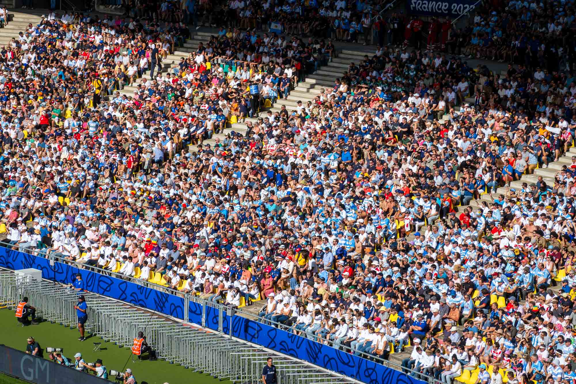 Tribune pleine de supporters argentins en ciel et blanc et japonais en rouge et blanc derrière une barrière bleue, photographes de presse au bord de la pelouse