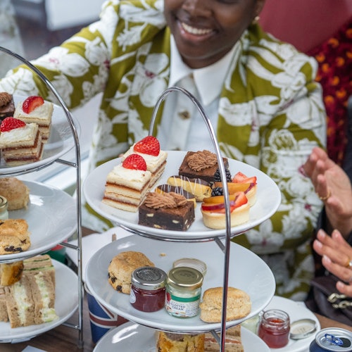 Personen genießen einen Nachmittagstee mit einem dreistöckigen Tablett voller verschiedener Desserts, Scones und Sandwiches. Ein lächelndes Gesicht ist teilweise sichtbar.
