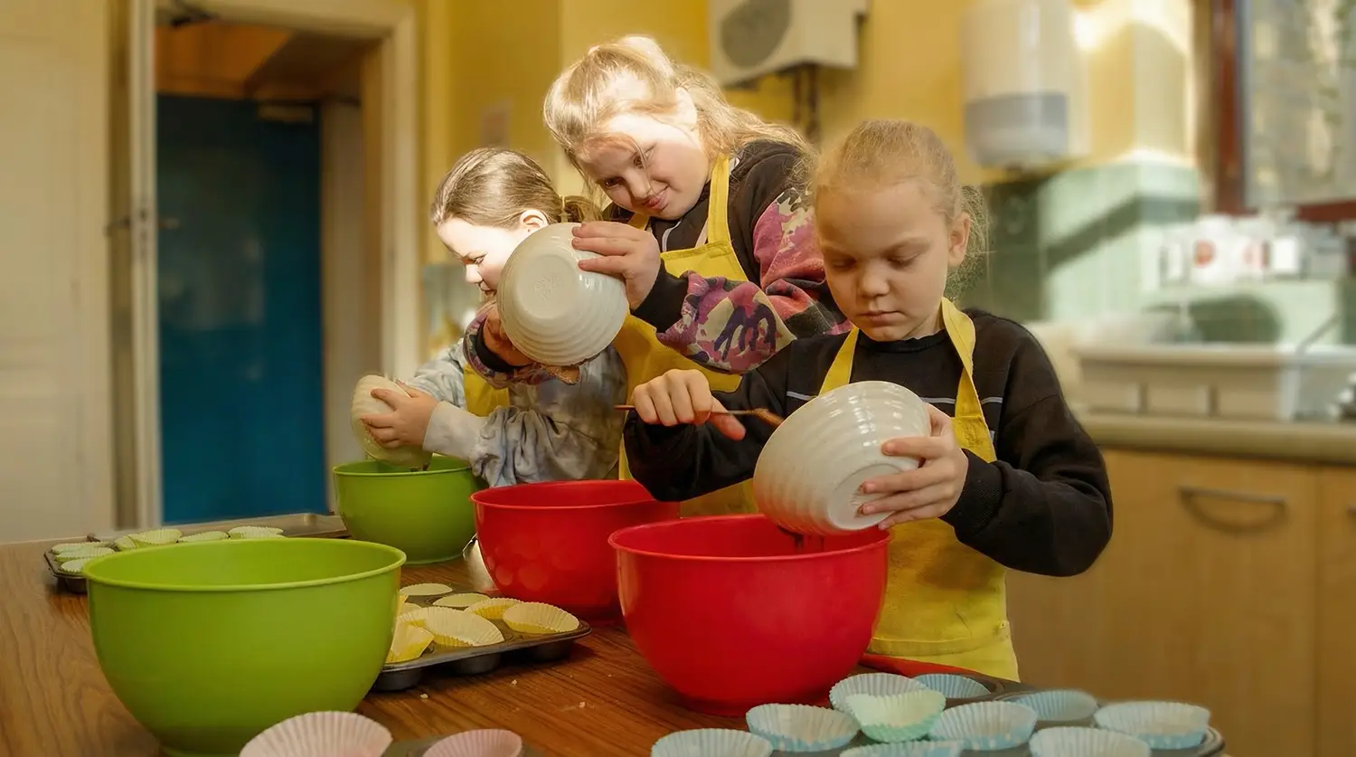 Children wearing aprons baking together at a table filled with bowls and cupcake cases.