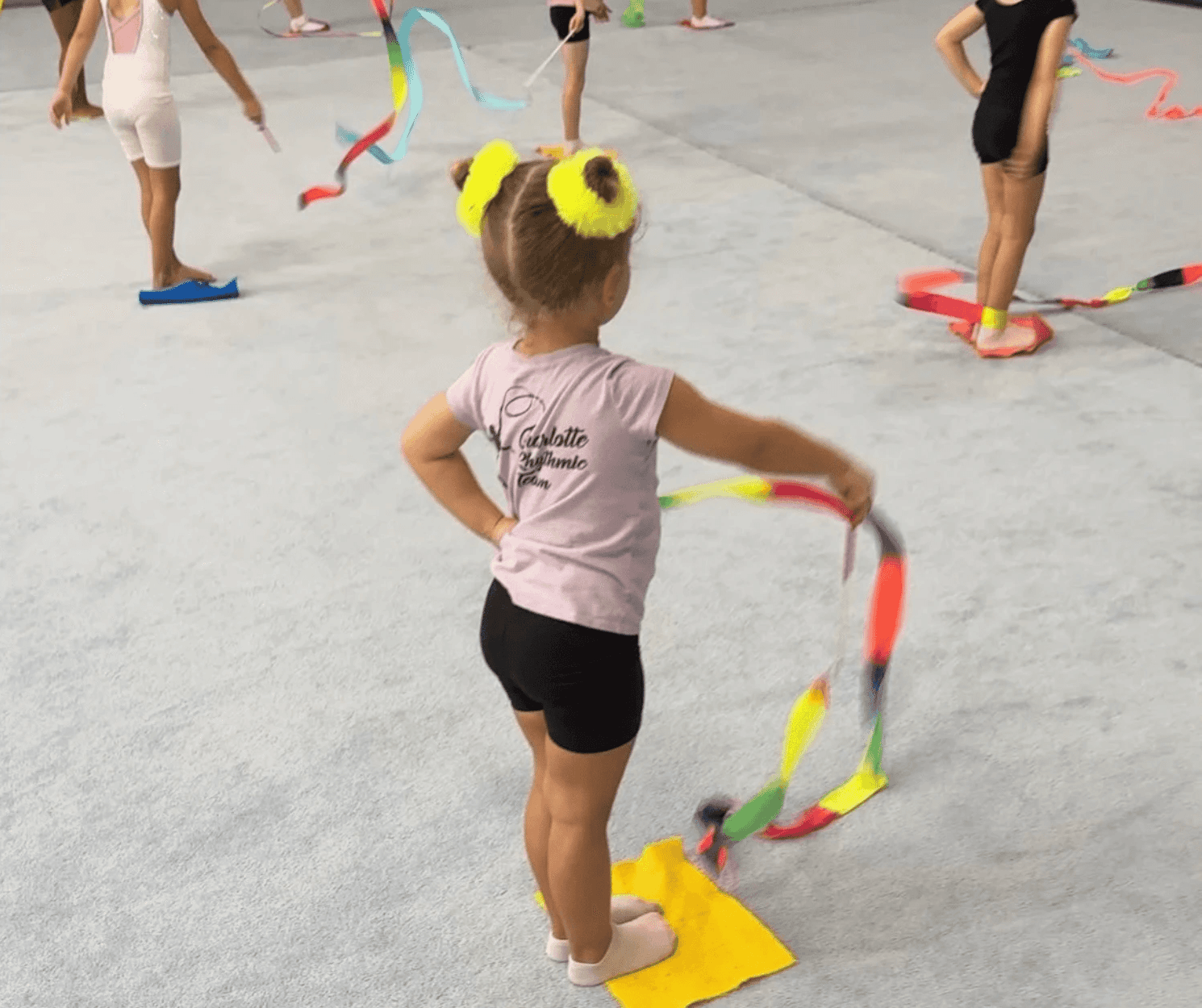 Adorable preschool girl with yellow pom-pom hair bows twirling a colorful rainbow ribbon during rhythmic gymnastics class at SportPlus Academy in Charlotte, building coordination and confidence in a fun preschool movement session.