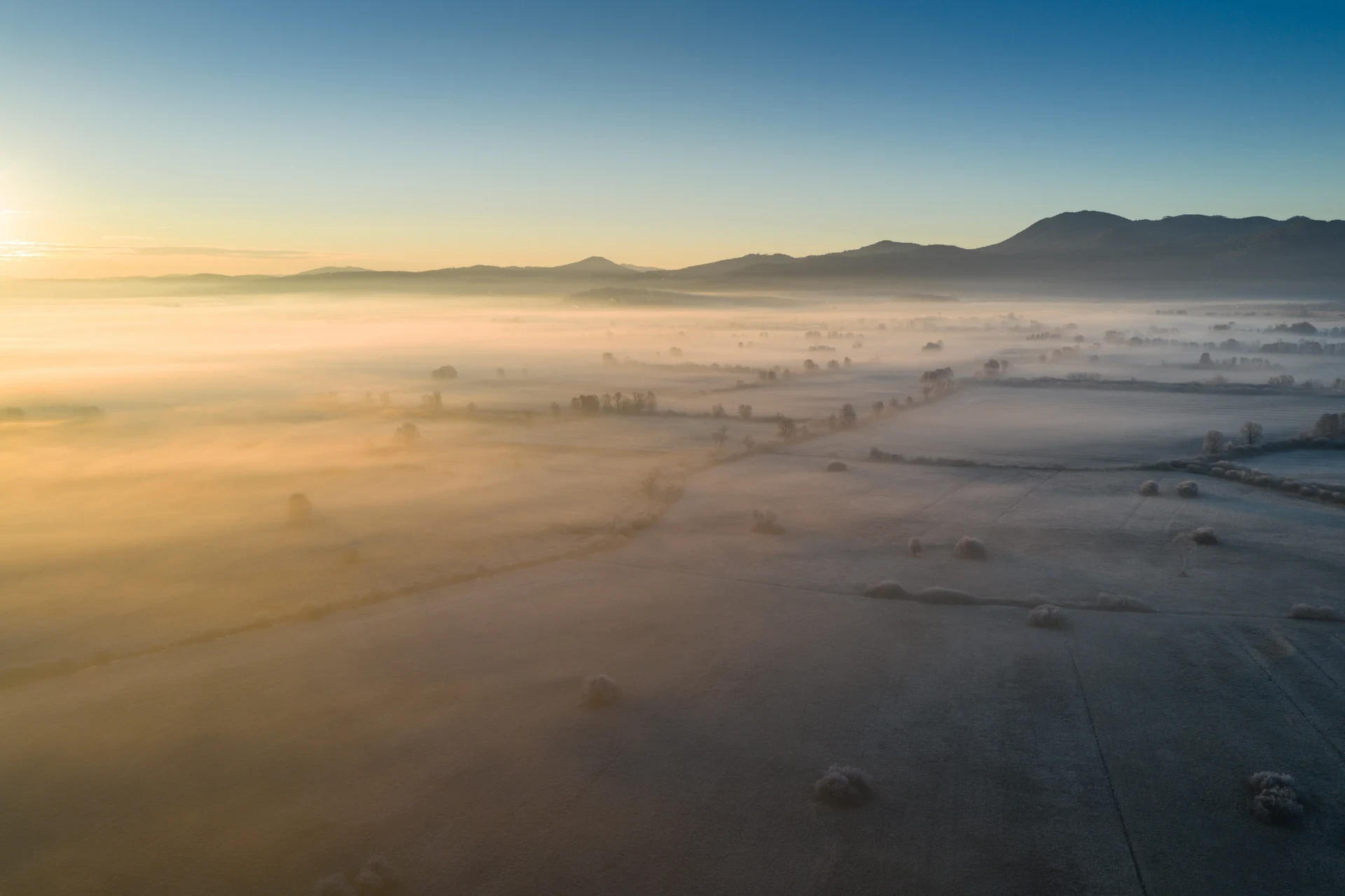  Aerial view of the Ljubljana Marshes in Slovenia at sunrise, featuring a thick layer of golden morning mist rolling over frost-covered fields and rural landscapes with distant hills under a clear blue sky.
