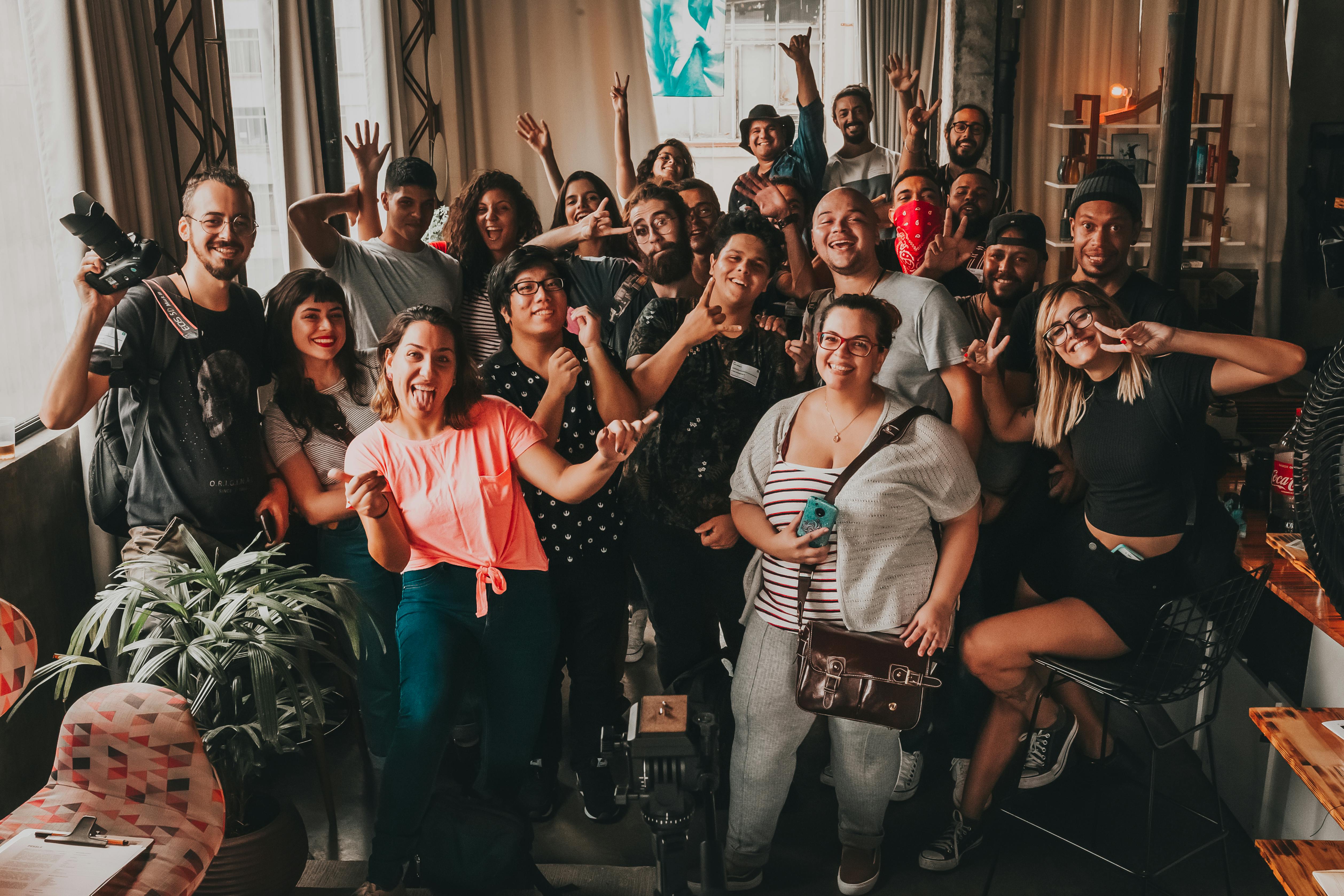 Large group of diverse, happy people posing indoors with hands up.
