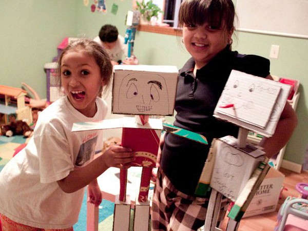 Two young girls stand side by side in a playroom. The girl on the left wears a white t-shirt and holds a red cardboard box with a face drawn on it. The girl on the right wears a black shirt and plaid skirt and holds a white cardboard box with a face drawn on it. A third child is visible in the background, partially obscured. Playroom toys and furniture are visible in the background.