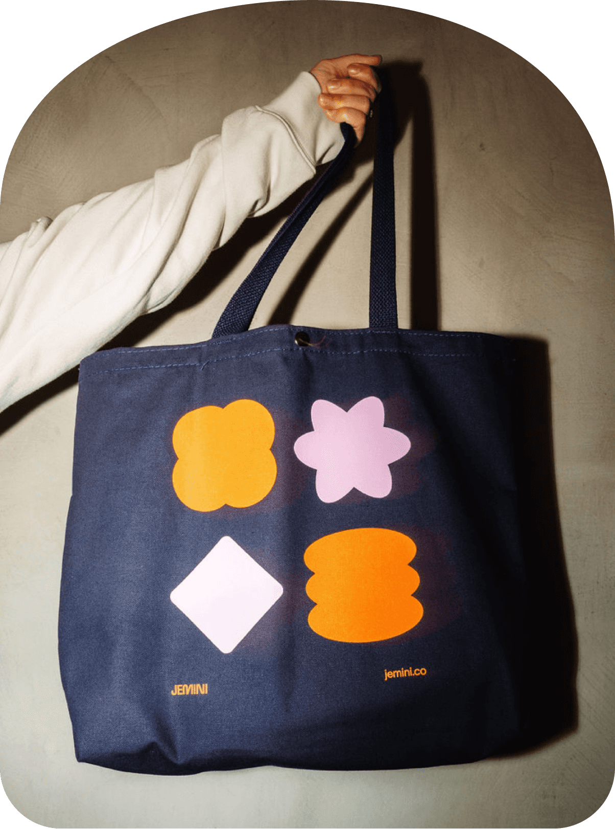Hand holding a navy tote bag featuring four colorful geometric shapes and small “jemini” branding near the bottom.