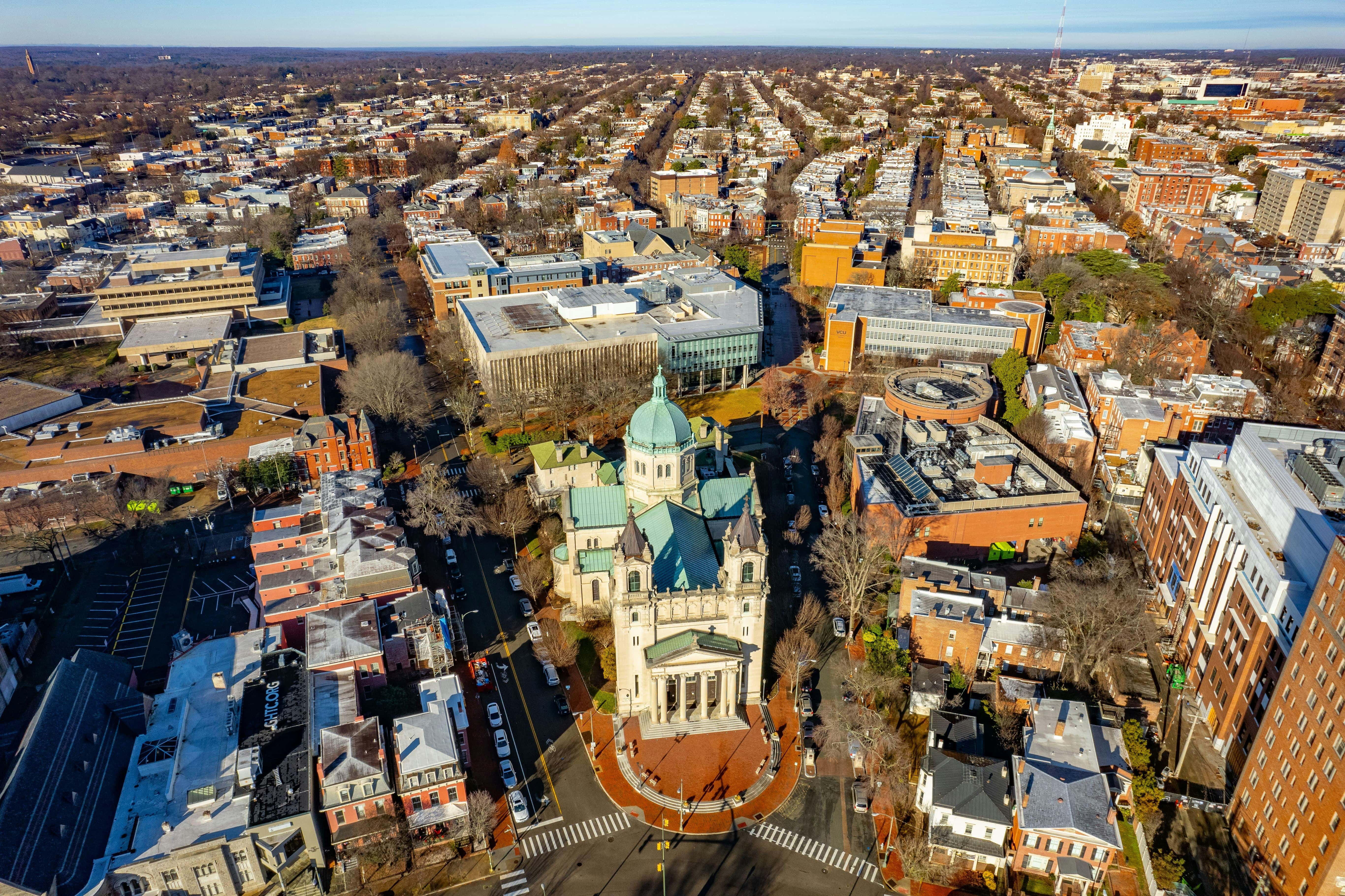 A wide aerial shot of a Virginia city, showcasing a historic church at the center, with surrounding residential and commercial buildings. The streets are lined with parked cars, and the city’s architectural blend of historic and modern structures is captured against a backdrop of tree-lined neighborhoods.