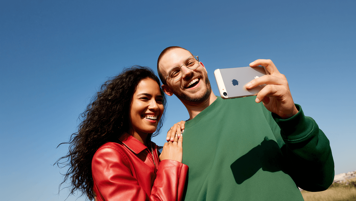 A smiling couple takes a selfie under a clear blue sky. The woman wears a red jacket, and the man, in a green top, holds a smartphone.