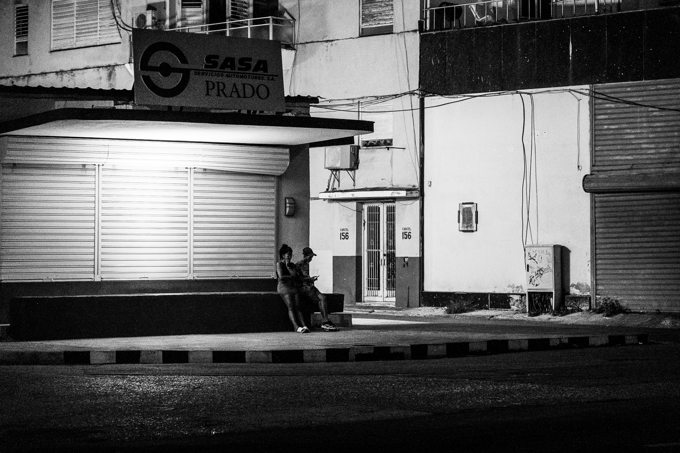 Black and white photograph of a couple standing by a closed shop on a quiet street, street photography by Richard Peterson.