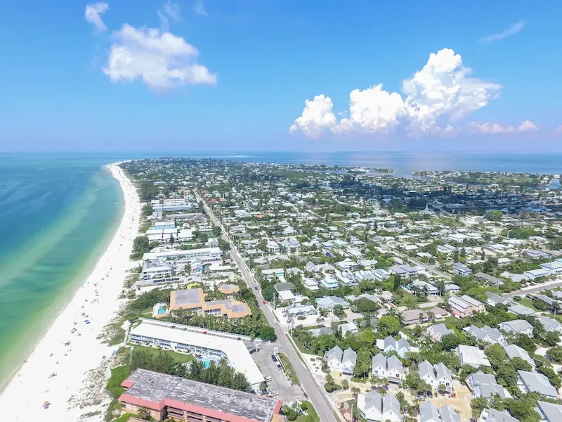 Aerial view of Anna Maria Island, FL, showcasing a stunning coastline with white sandy beaches and turquoise waters, alongside a charming residential area under a bright blue sky with scattered clouds.