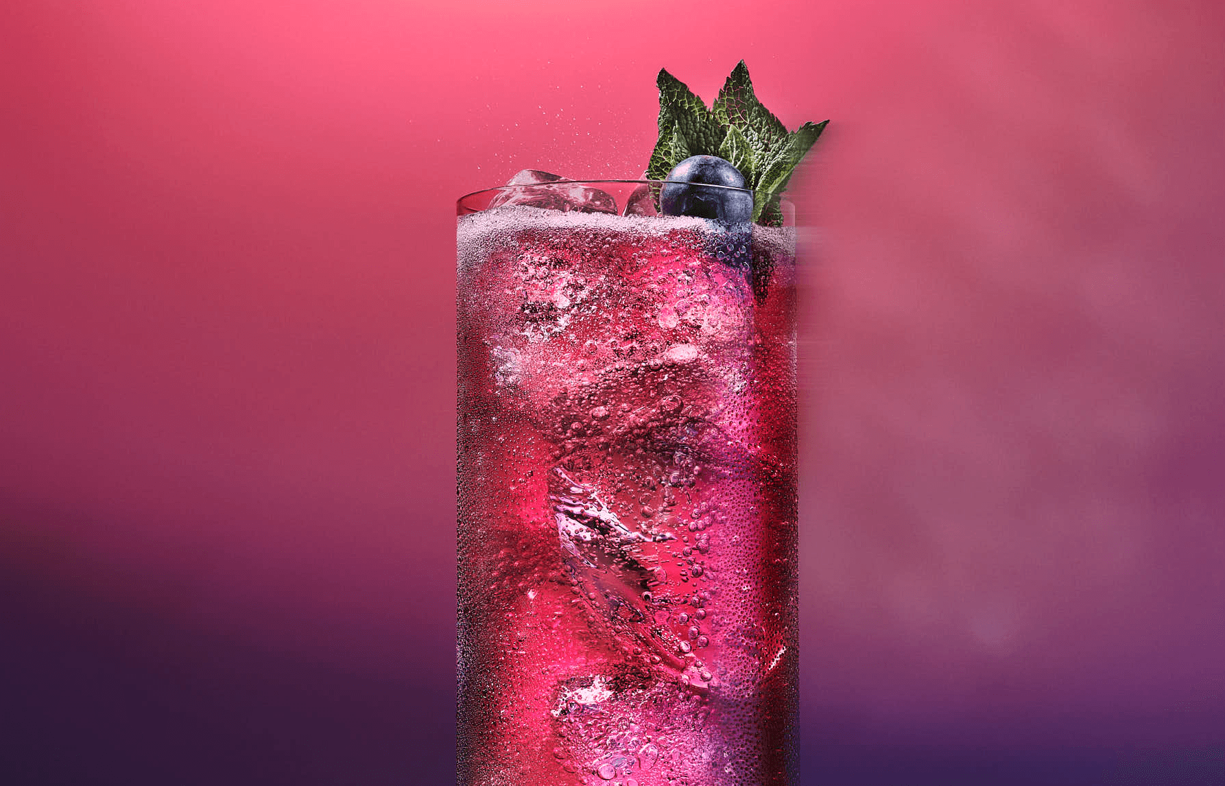 A vibrant pink drink with ice, garnished with mint leaves, against a soft purple background.