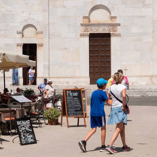 People walking in front of an outdoor café with an ornate menu board and a large building with arches and a wooden door in the background.