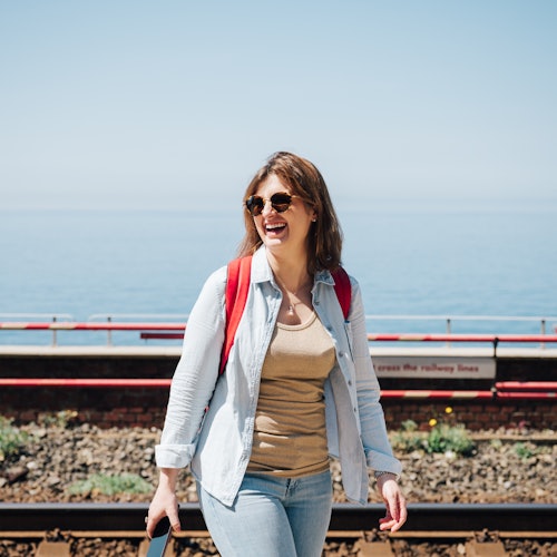 Žena s červeným batohem a slunečními brýlemi kráčí poblíž železničních tratí s výhledem na moře v pozadí.