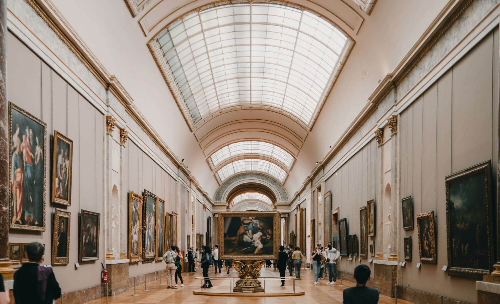 A grand museum hallway with a high arched ceiling, adorned with classical paintings on both sides. Visitors admire the art, creating a serene atmosphere.