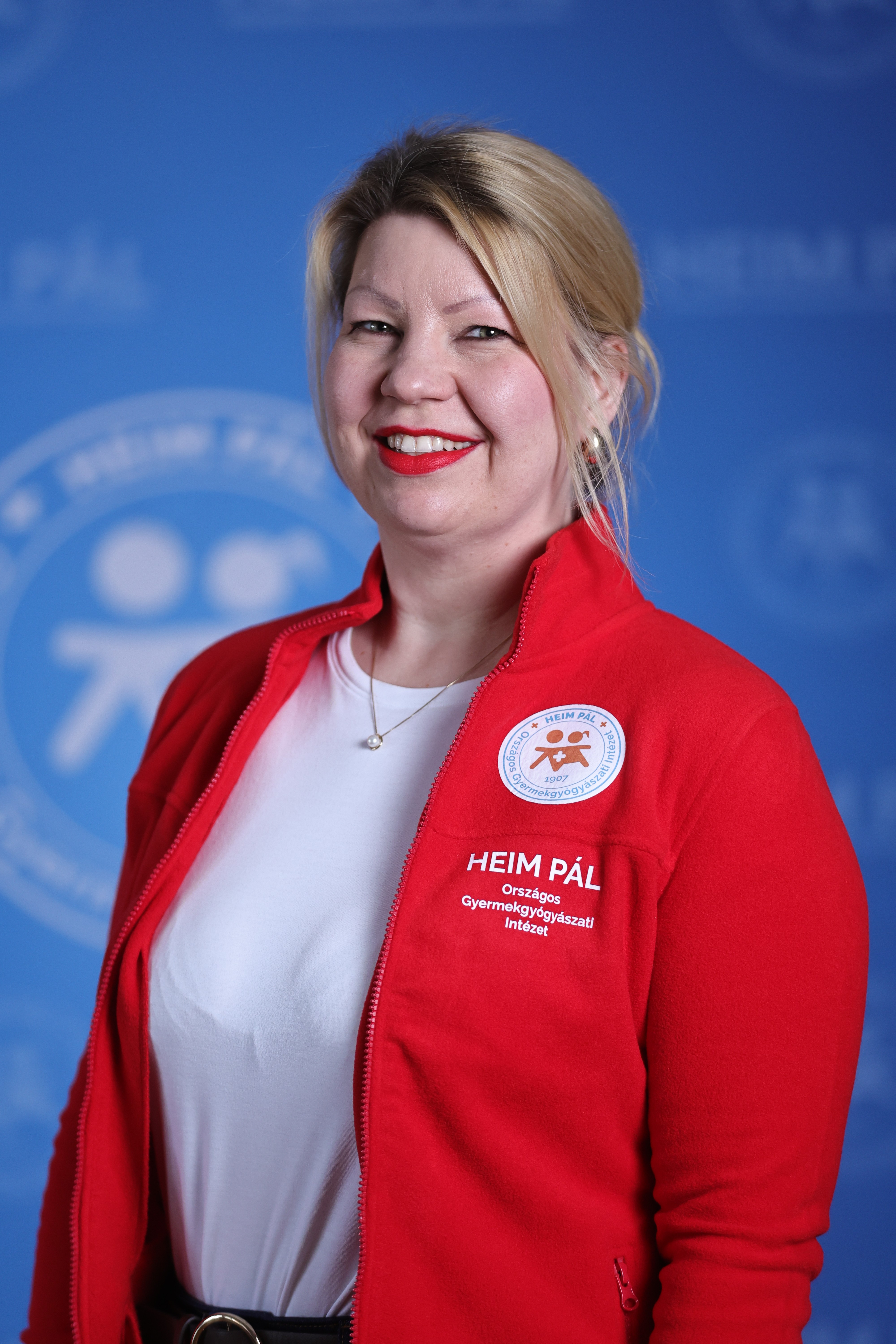 Szőke hajú nő, Heim Pál feliratos piros pulóverben kék háttér előtt.