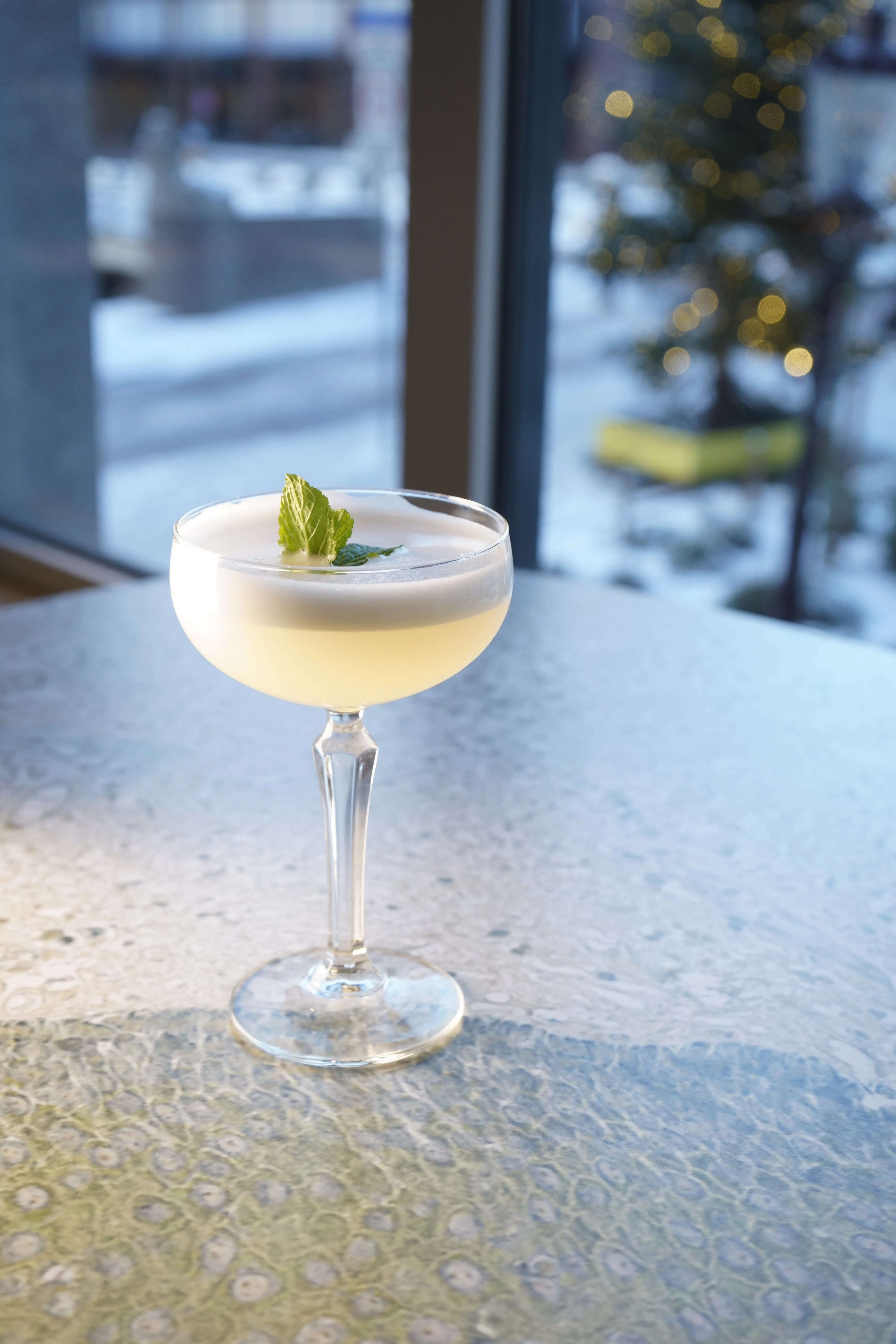 A chilled cocktail in a coupe glass garnished with a lime slice, set on a table with a blurred background.