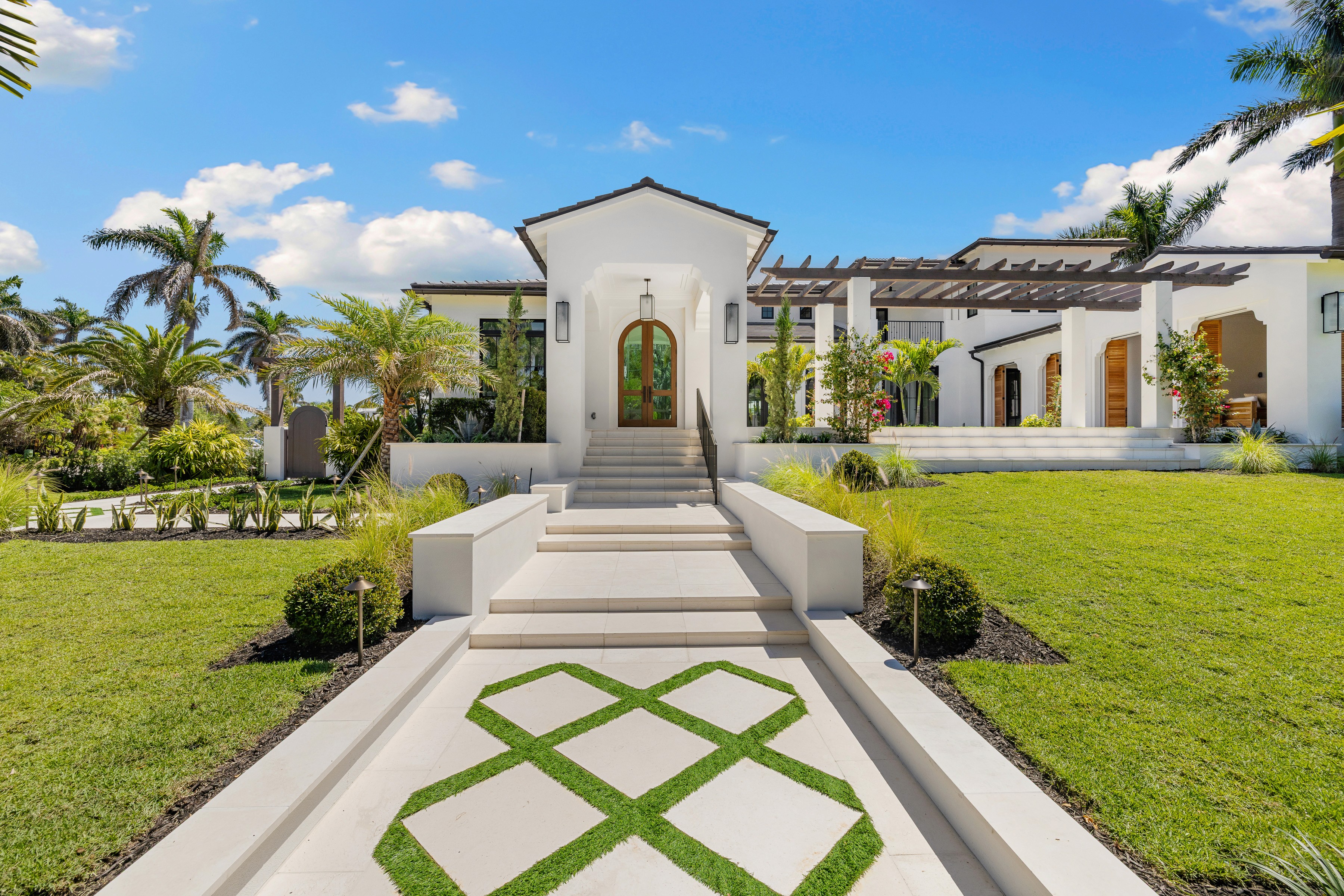 a luxurious walkway leading up to a white modern home with palm trees and vibrant green grass on both sides