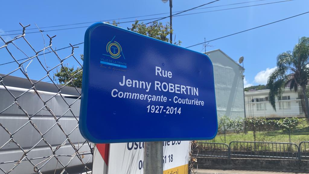 Une plaque bleue indique en français « Rue Jenny Robertin, Commerçante - Couturière, 1927-2014 ». Elle est fixée à côté d'une clôture grillagée sous un ciel bleu clair.