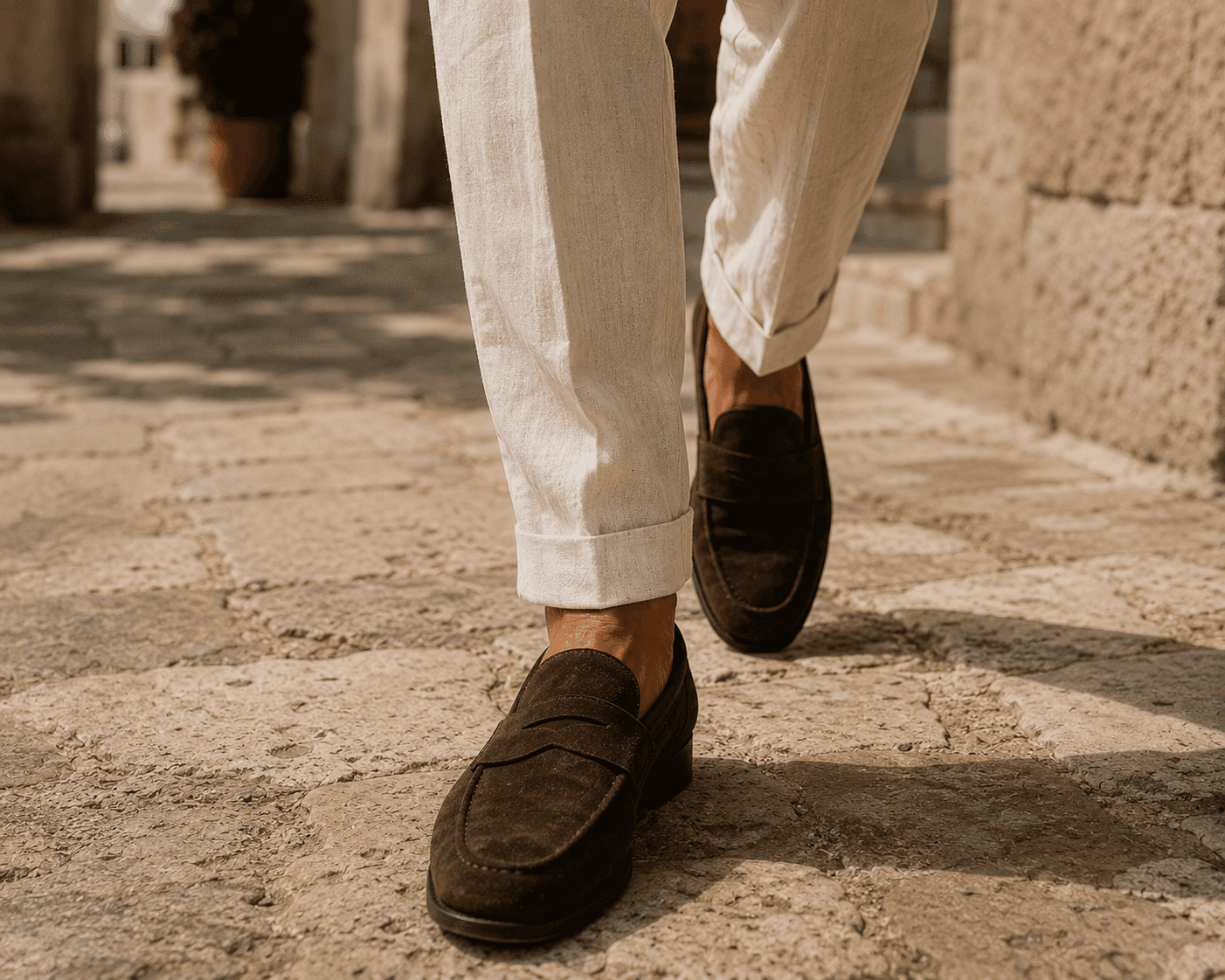 A close-up, ground-level shot of a man's feet walking forward on a rustic, sunlit stone-paved path. He is wearing classic dark brown suede penny loafers without socks, paired with light cream-colored linen trousers featuring distinct cuffed hems and sharp front creases. The background is softly blurred, showing glimpses of stone structures and a terracotta pot.
