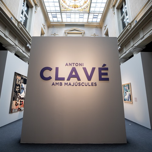 Art gallery na may malaking sentral na display na nagbabasa ng "Antoni Clavé Amb Majúscules." Ang mga nakapaligid na pader ay nagtatampok ng mga naka-frame na obra ng sining.