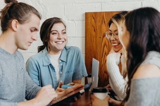 A group of 4 friends sitting at a table in a coffee shop. One man is looking at an iPad and the other 3 ladies are chatting and smiling.