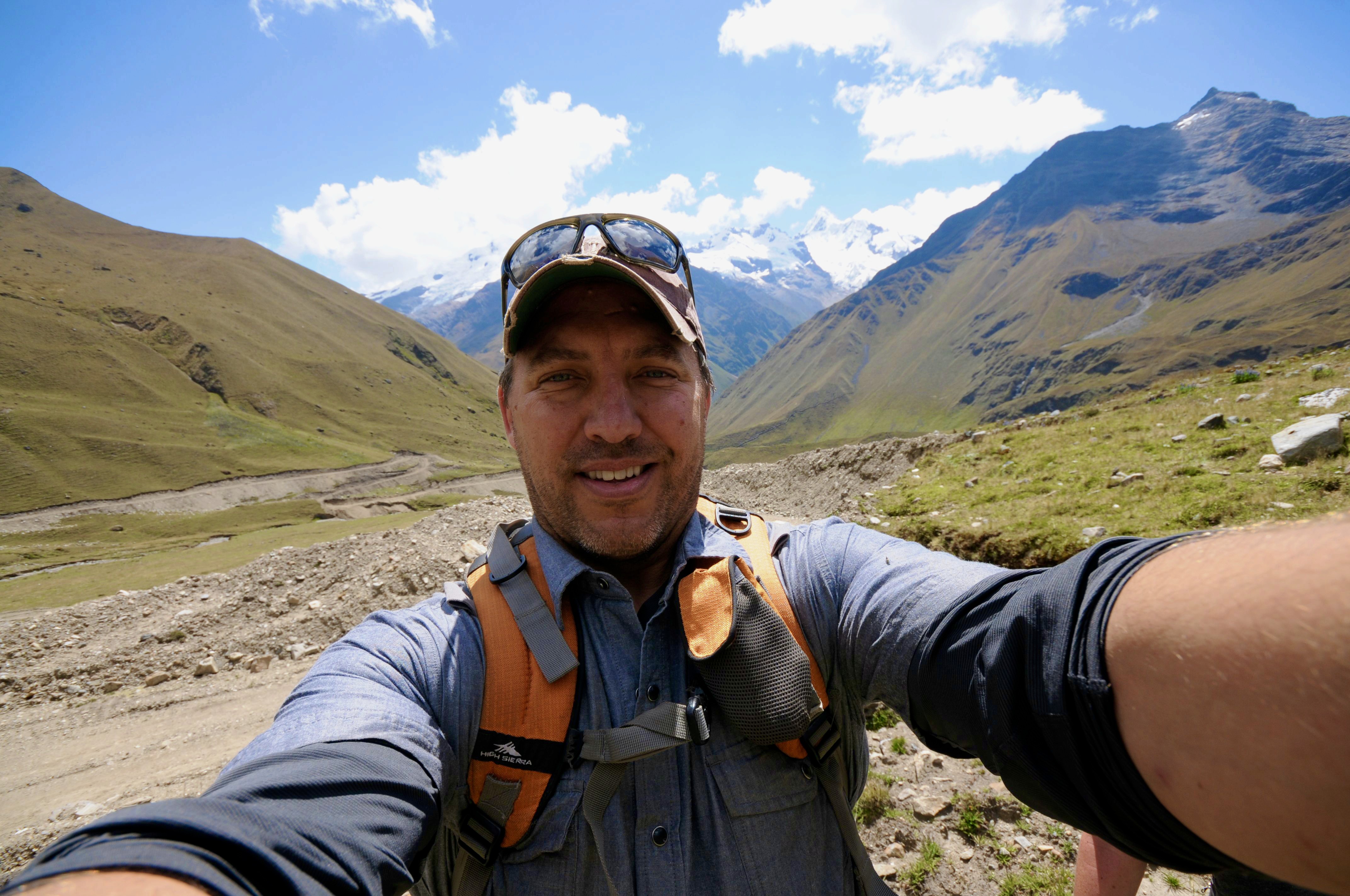 Director Ross Fenter, known for his work in content and documentary, takes a selfie while hiking in a remote mountain range. Wearing a cap, sunglasses, and a backpack, he smiles against a backdrop of rugged trails, rolling hills, and snow-capped peaks under a clear blue sky.
