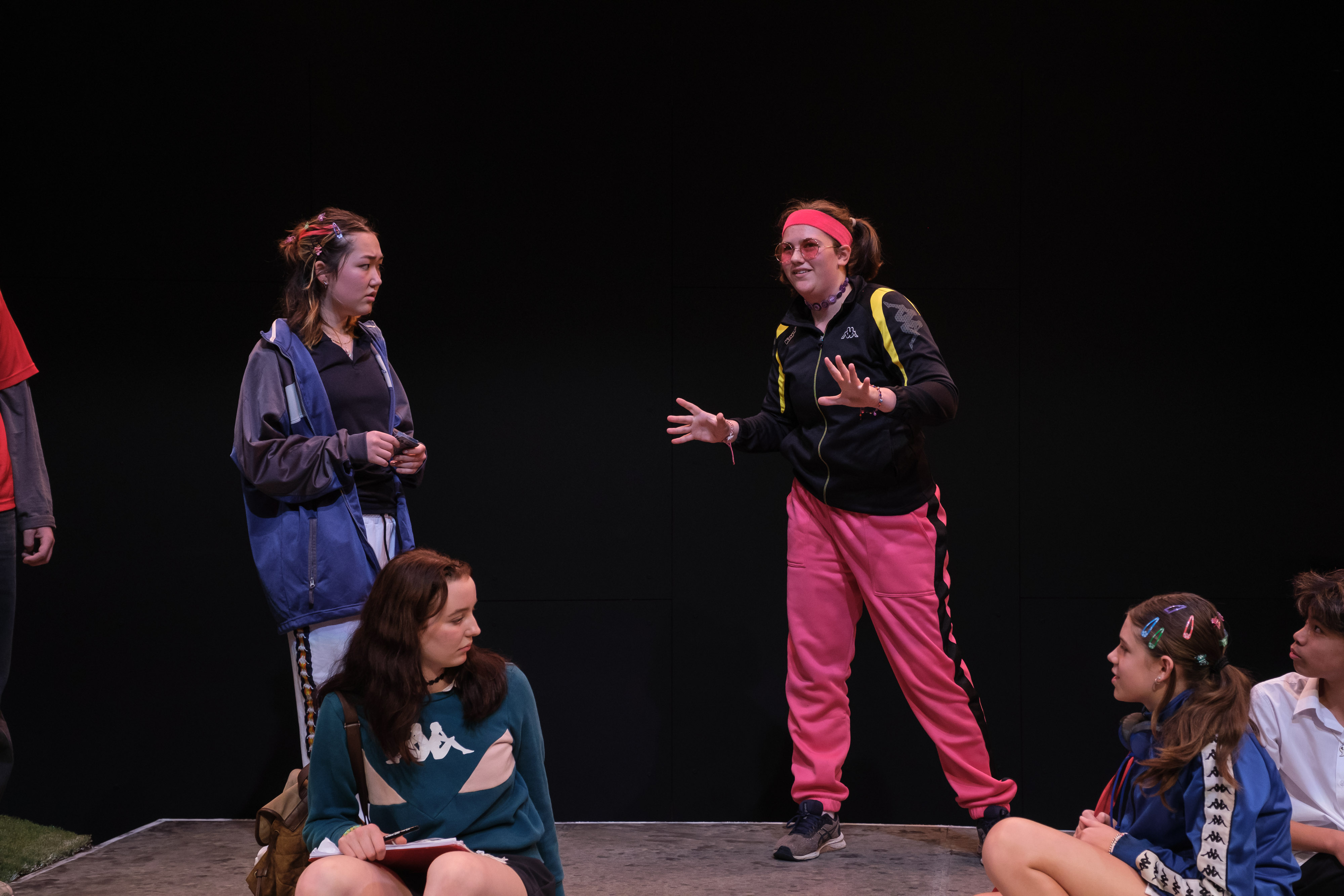 A cast of teenagers performs an energetic scene. One performer in bright pink track pants speaks animatedly to a seated group, while others watch with confused or curious expressions, set against a minimalist school backdrop.