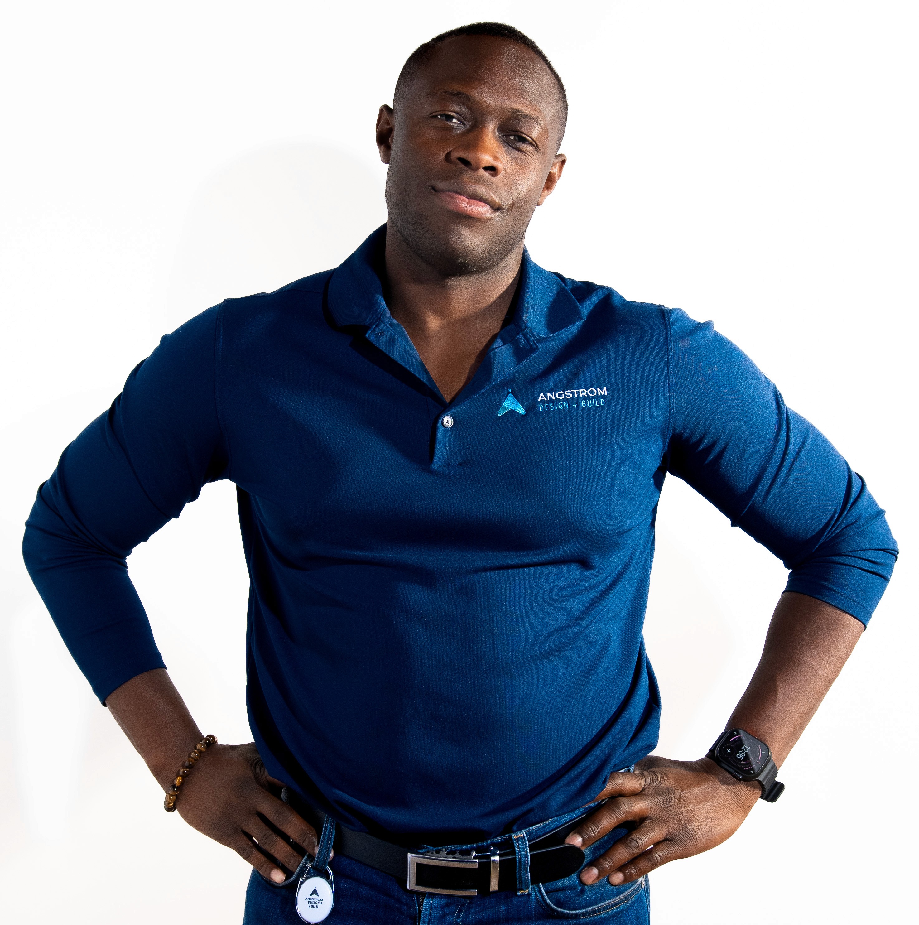 A smiling person with curly hair sits at a desk, looking at a laptop against a dark background.