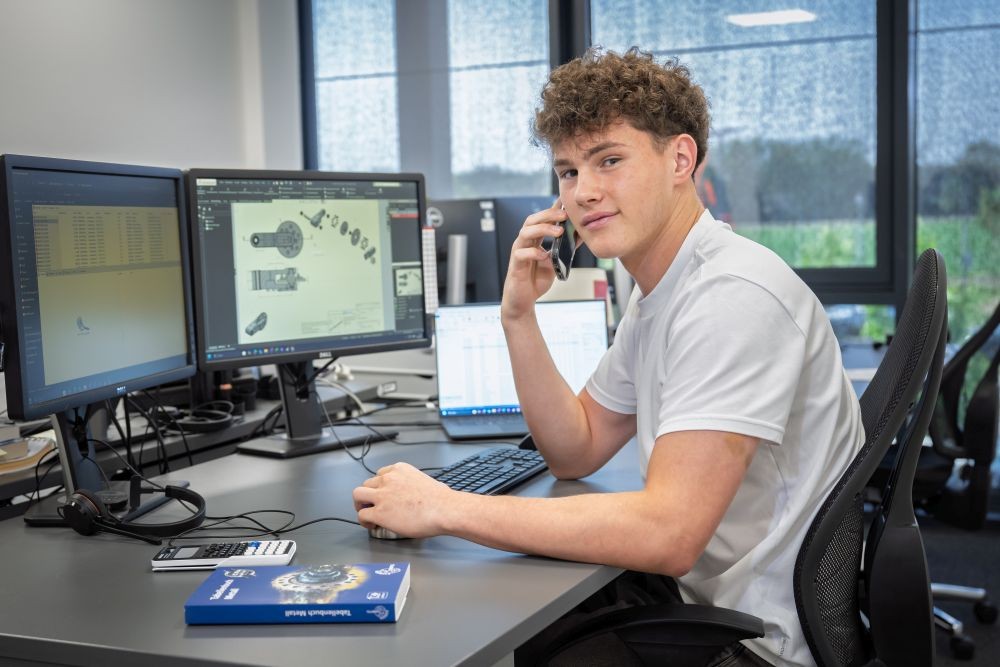 Ein junger Mann mit lockigem Haar lächelt, während er am Telefon spricht. Er sitzt an einem Schreibtisch mit zwei Monitoren und einem Laptop.