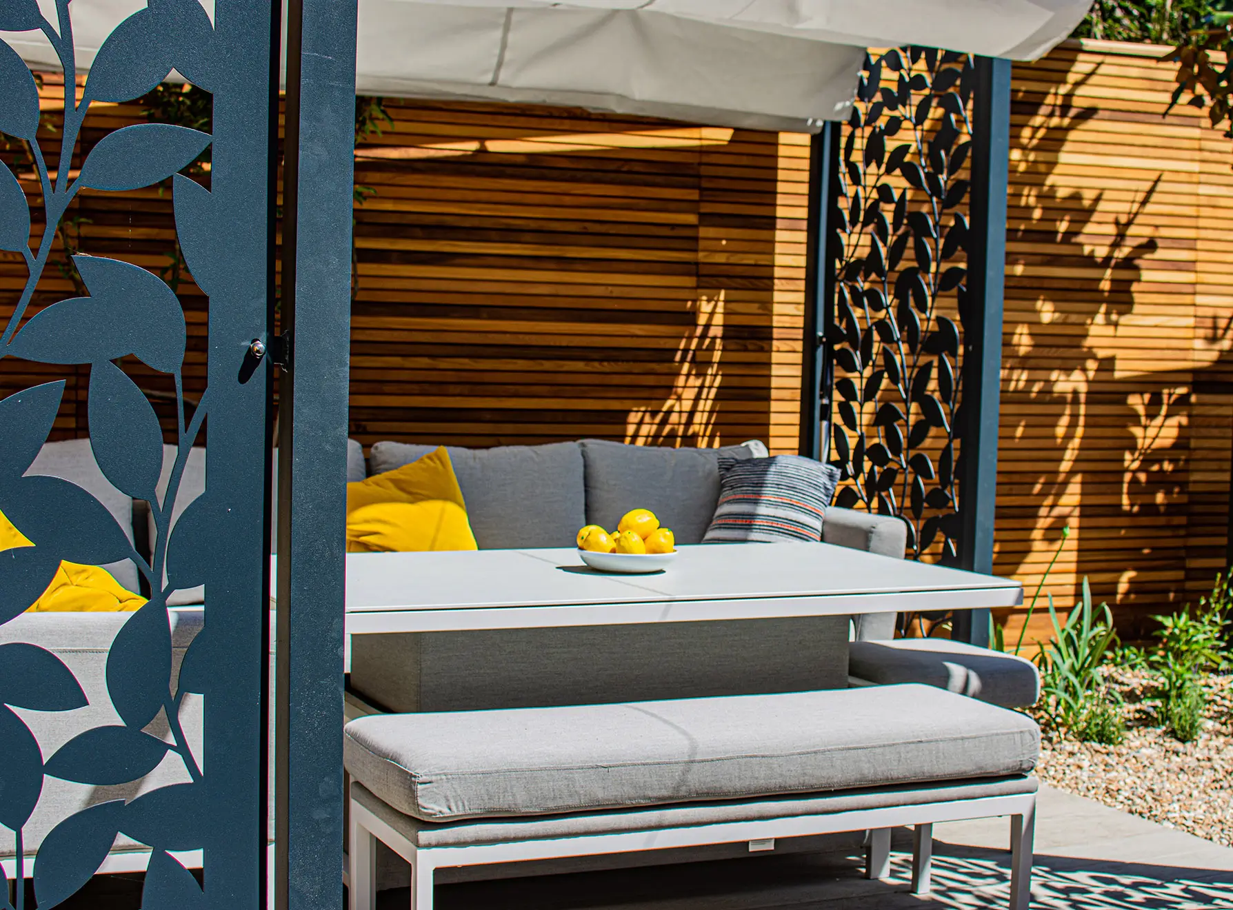 A cozy outdoor seating area with a gray sofa and bright yellow cushions, surrounded by greenery.