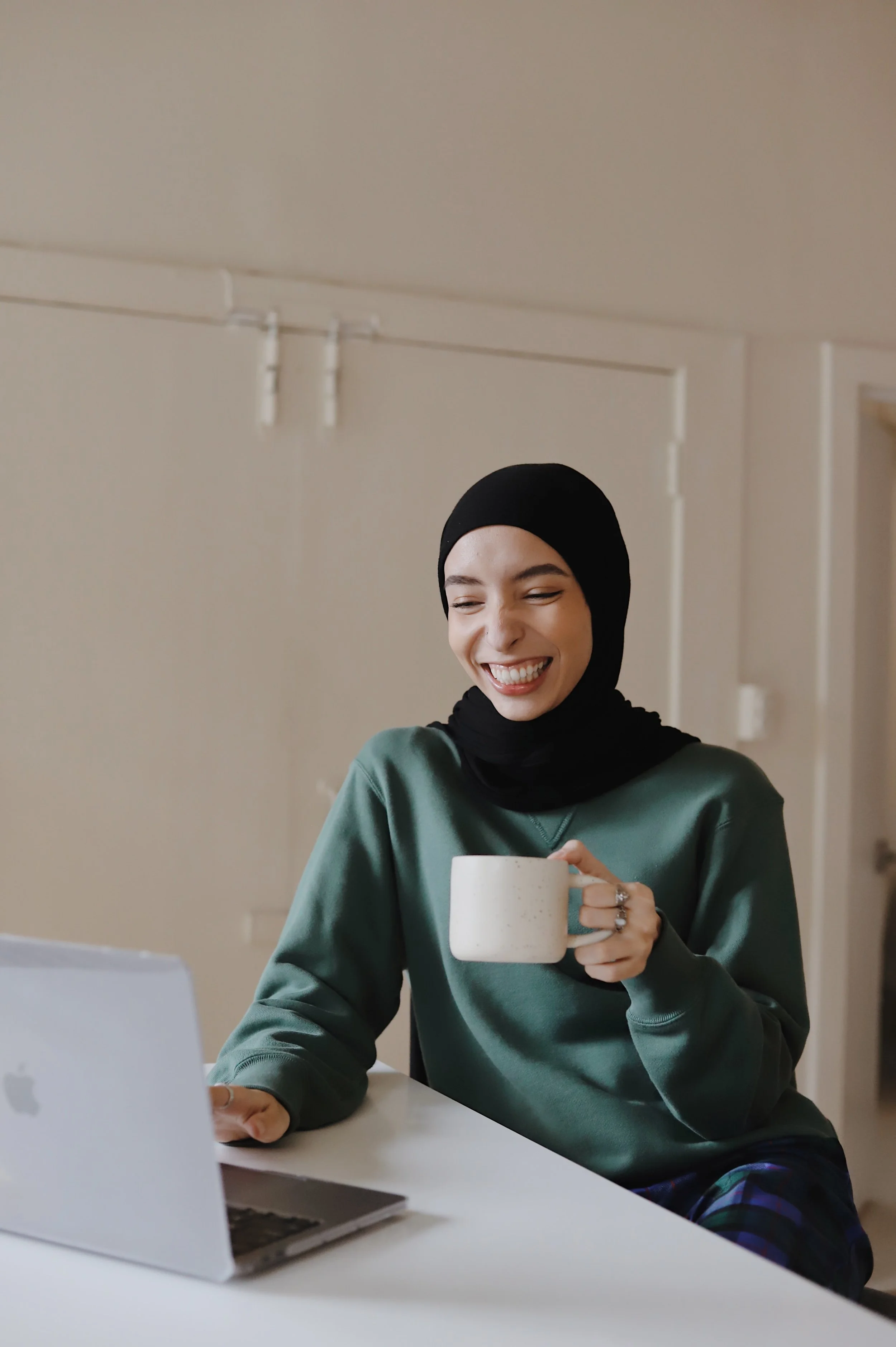 A woman in a hijab smiles while having a cup of tea at her laptop