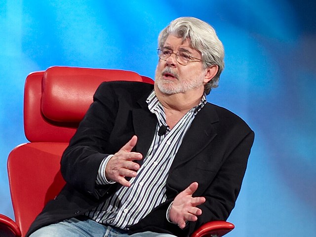 George Lucas conducting an interview, he is sat in a red chair, the set back ground is blue