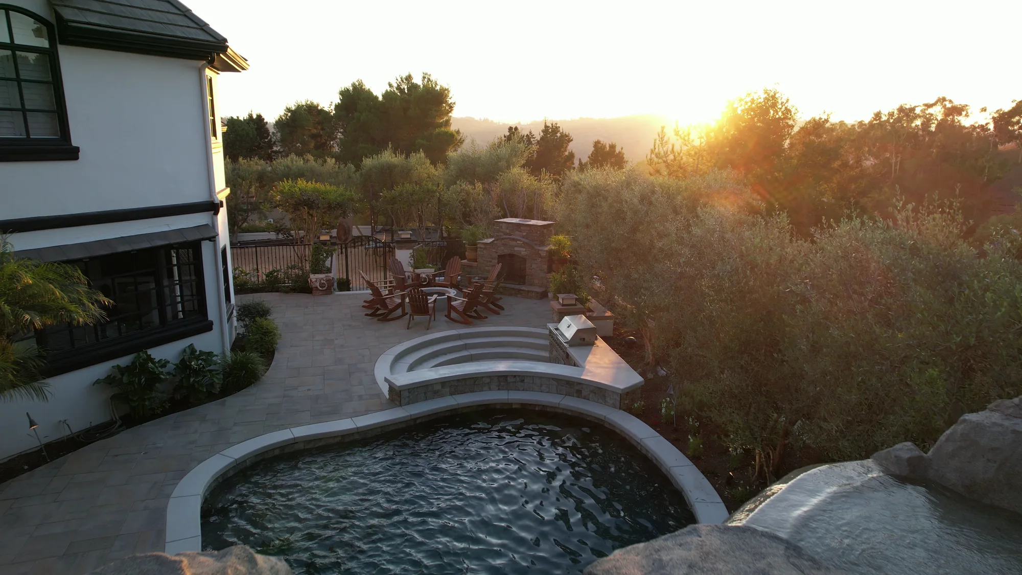 View from above the rock grotto toward the curved pool, terrace steps and outdoor seating at sunset.