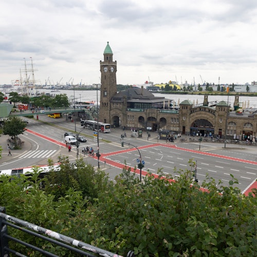 A clock tower and brick building next to a busy intersection overlook a harbor with cranes and ships under a cloudy sky.